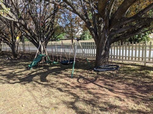Shady backyard with large trees, a white picket fence, and a green swing set on dry ground.