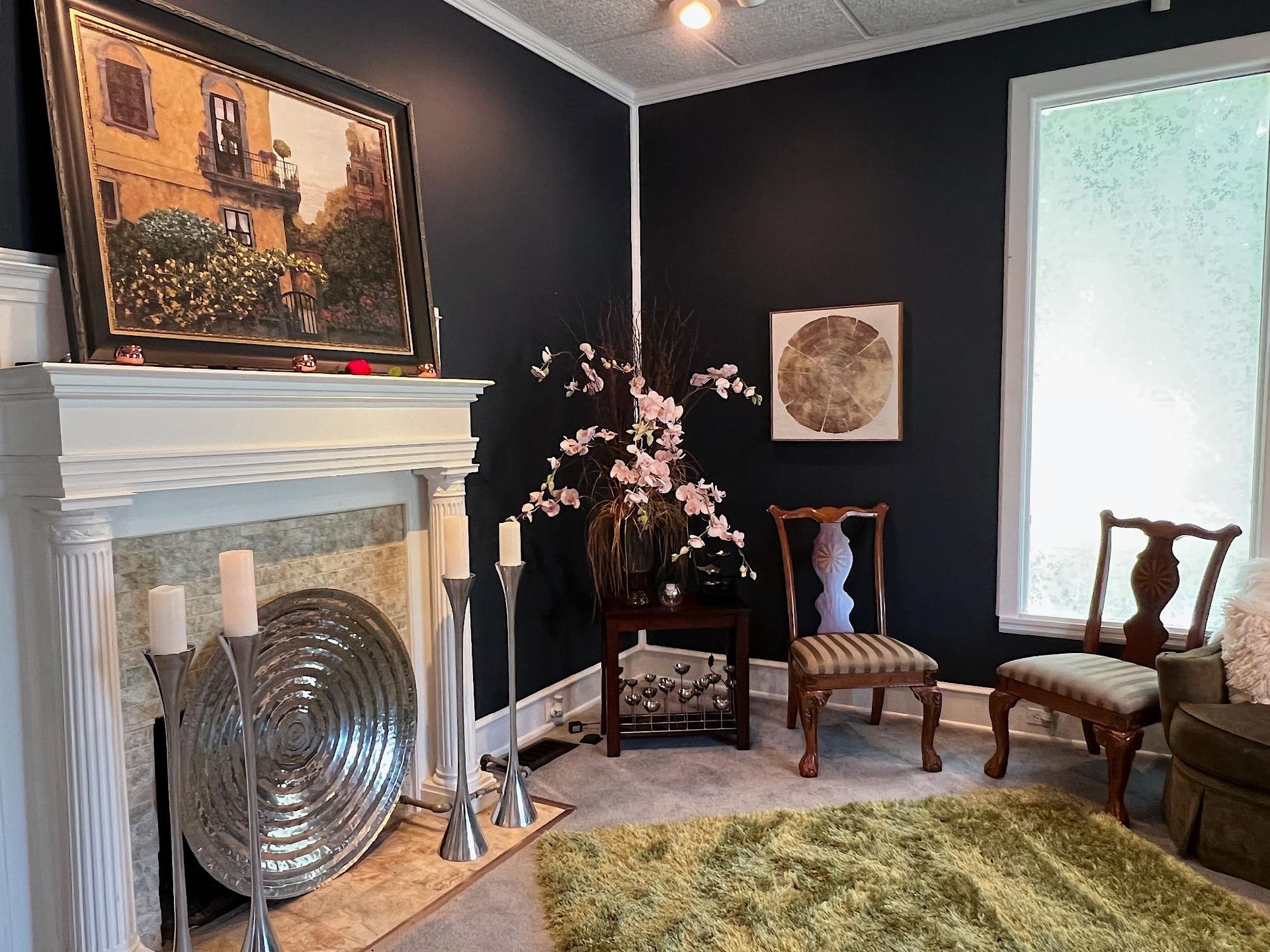 Elegant living room with black walls, white fireplace, vintage chairs, mirror, and large floor fan in front.