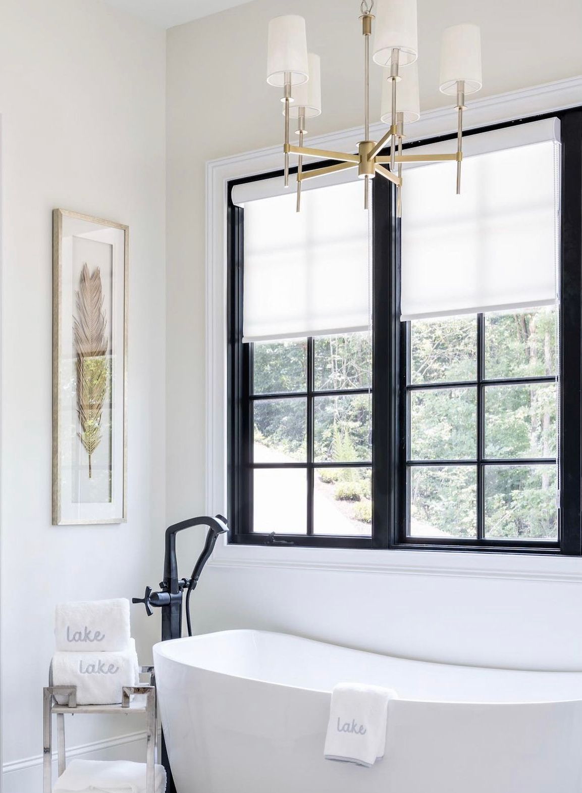 A bathroom with a tub , two windows and roller shades.