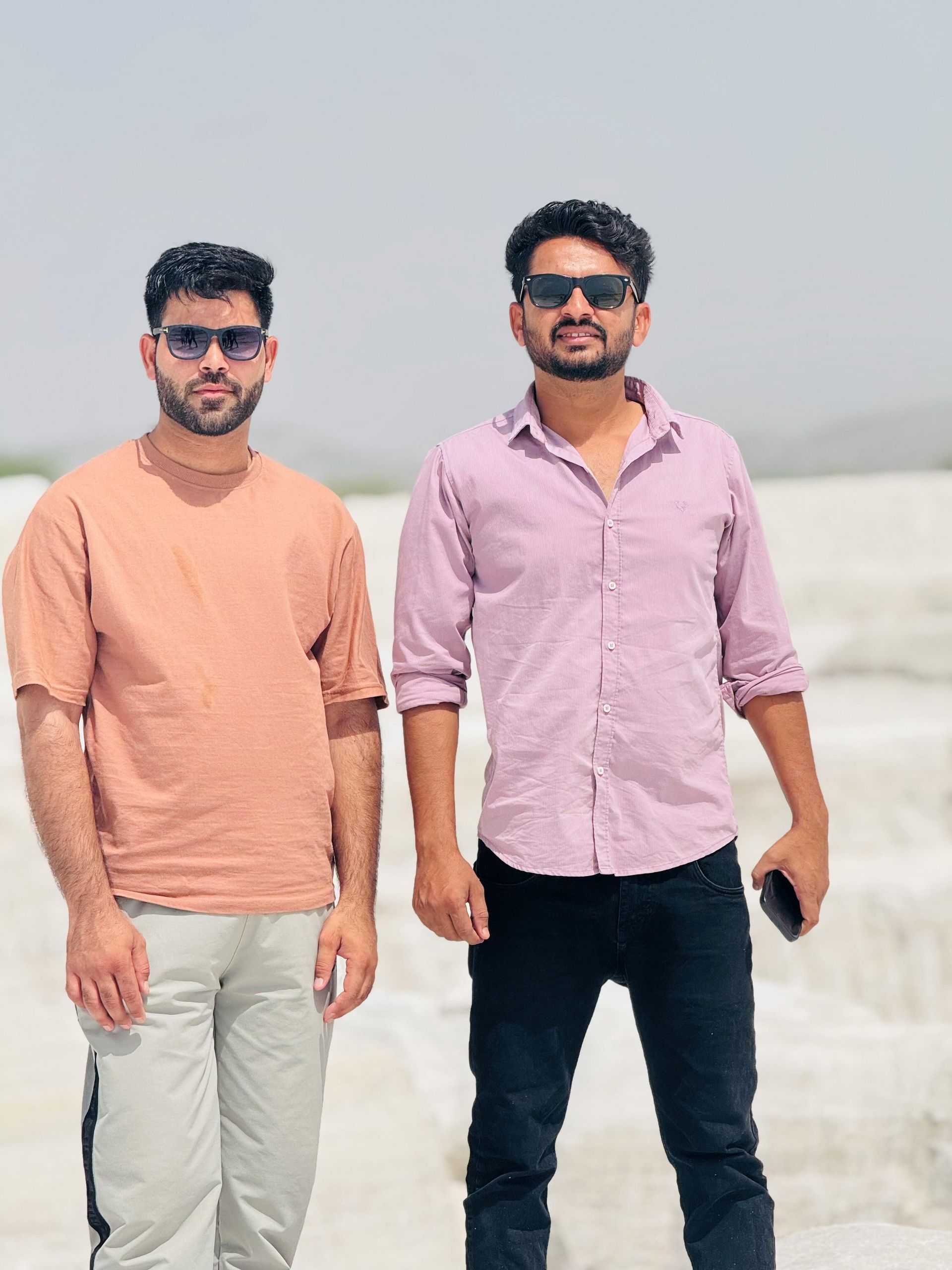 Two men in sunglasses standing outdoors, one in a peach shirt and the other in a pink shirt, posing for a photo.