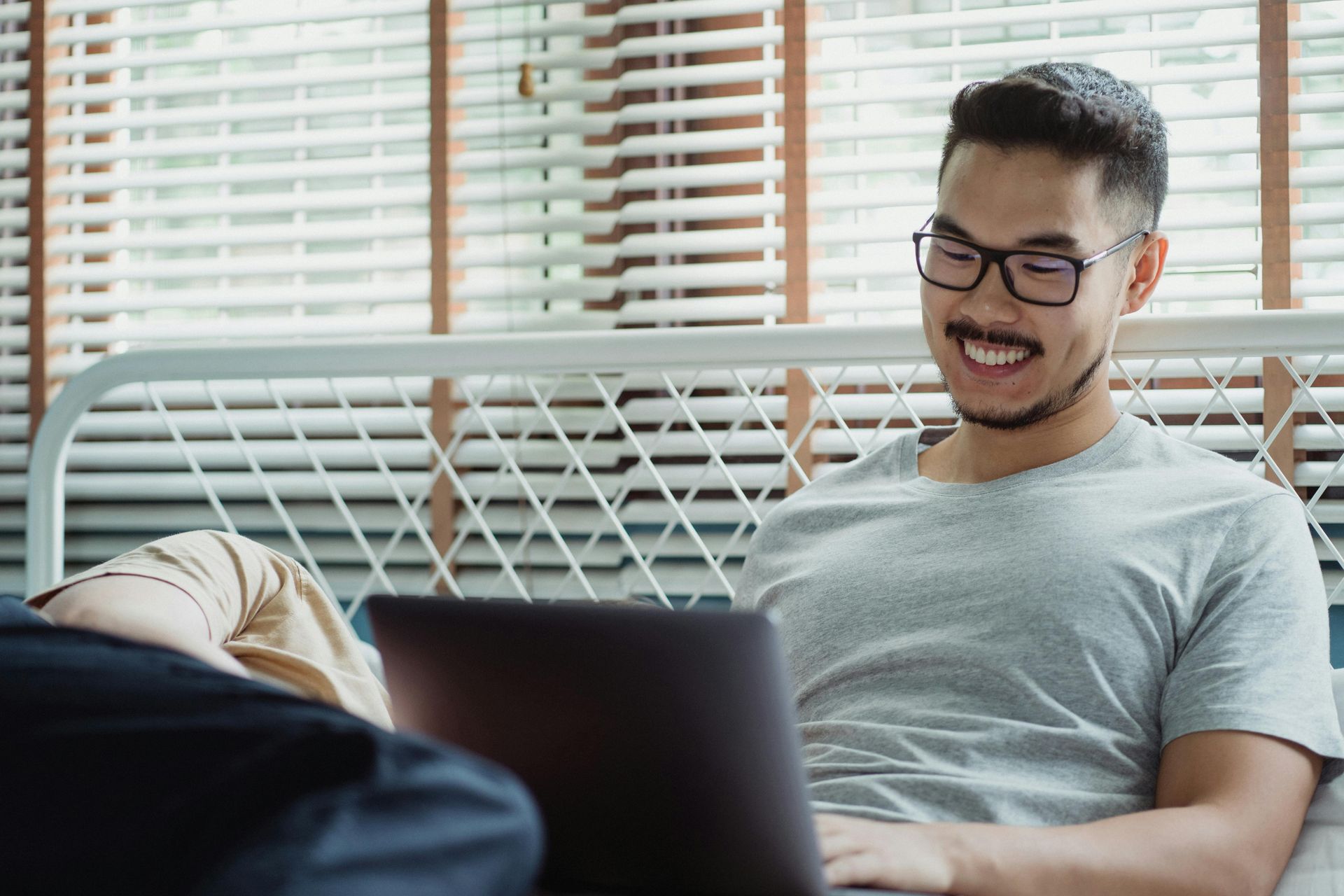 Man with glasses smiles while using a laptop in bed, near a window with blinds.