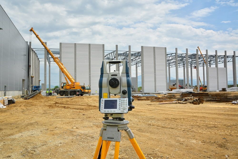 A Total Station Is Sitting On A Tripod In Front Of A Large Building Under Construction — CalCo Surveyors Pty Ltd in Gloucester, NSW