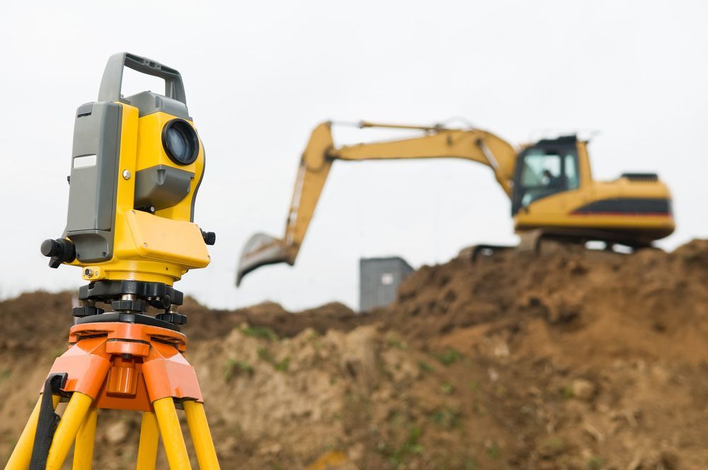 A Construction Site With A Yellow Excavator In The Background And A Theodolite In The Foreground — CalCo Surveyors Pty Ltd in Wingham, NSW