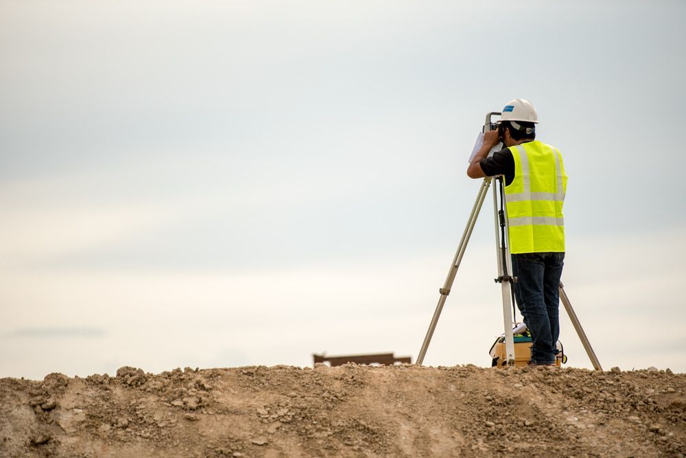 A Man Is Standing On Top Of A Dirt Hill Looking Through A Telescope — CalCo Surveyors Pty Ltd in Forster, NSW