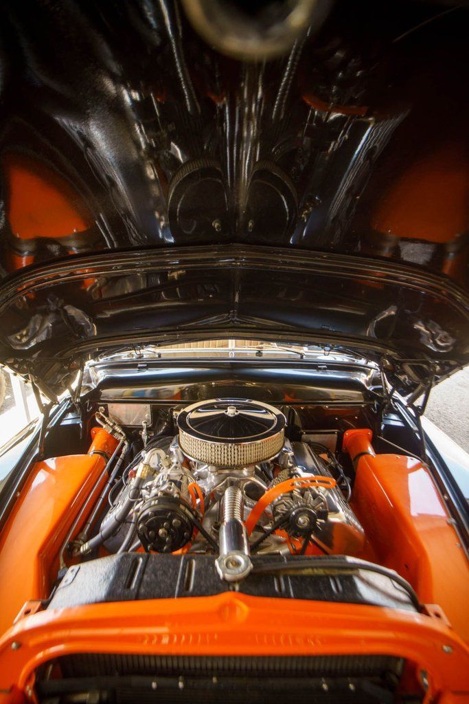 A high-angle view into the open hood of a classic car with a detailed, chrome engine and bright orange wheel wells.