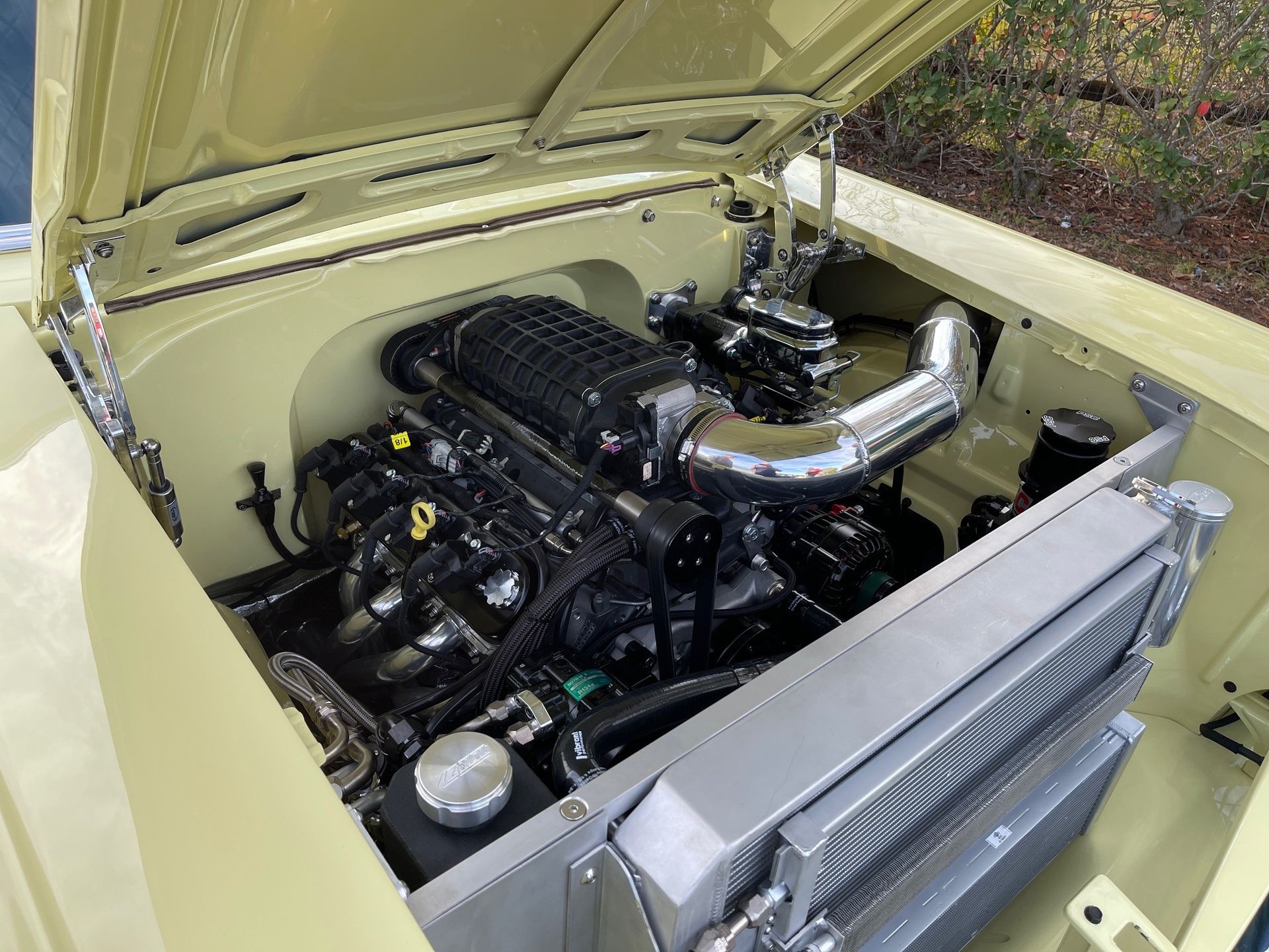 A high-performance supercharged engine with a polished intake pipe installed in a pale yellow classic car engine bay.