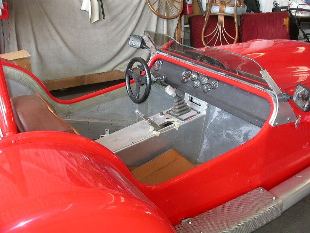 Interior of a red, open-top sports car, showing the steering wheel, gauges, gear shifter, and bucket seat in a garage.