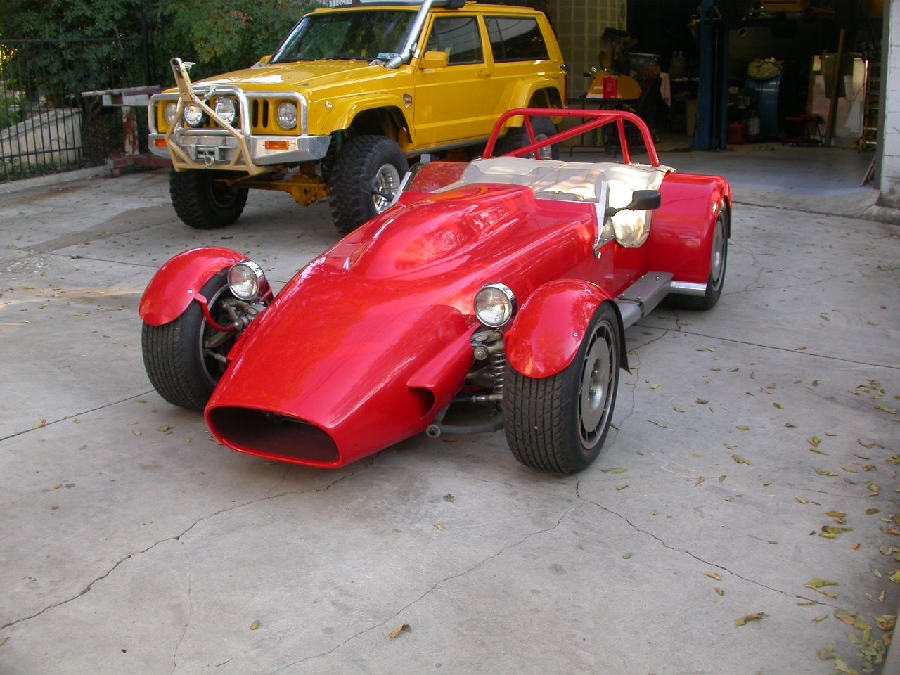 A bright red, open-wheel kit car parked in a driveway next to a yellow SUV.