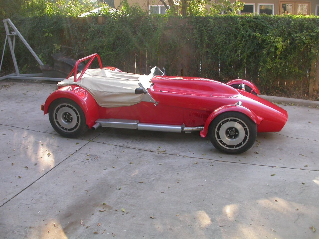 A bright red sports car with a beige tonneau cover parked on a concrete driveway in front of a hedge.
