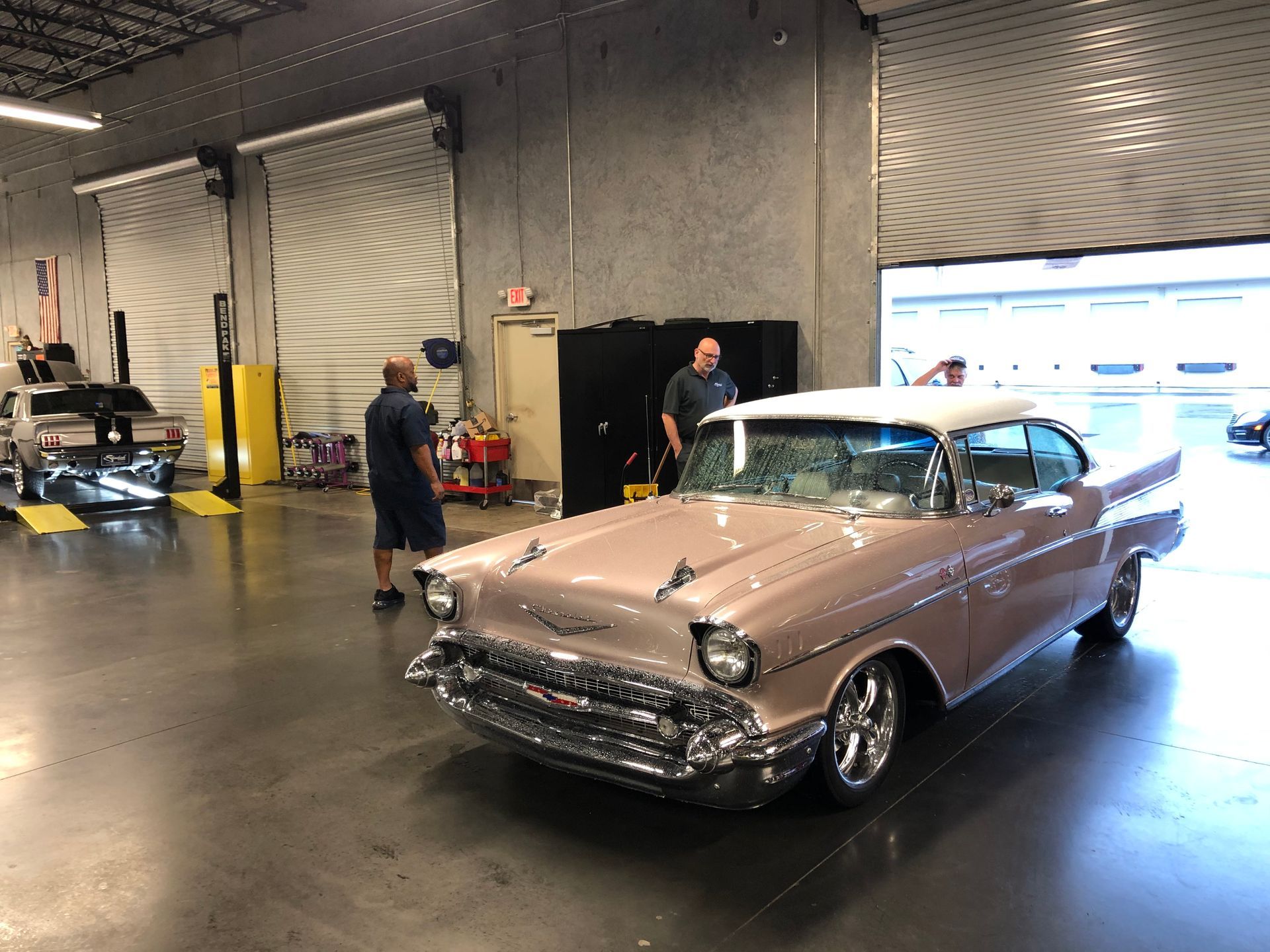 A dusty rose and white classic car parked in a garage workshop with two people standing nearby.