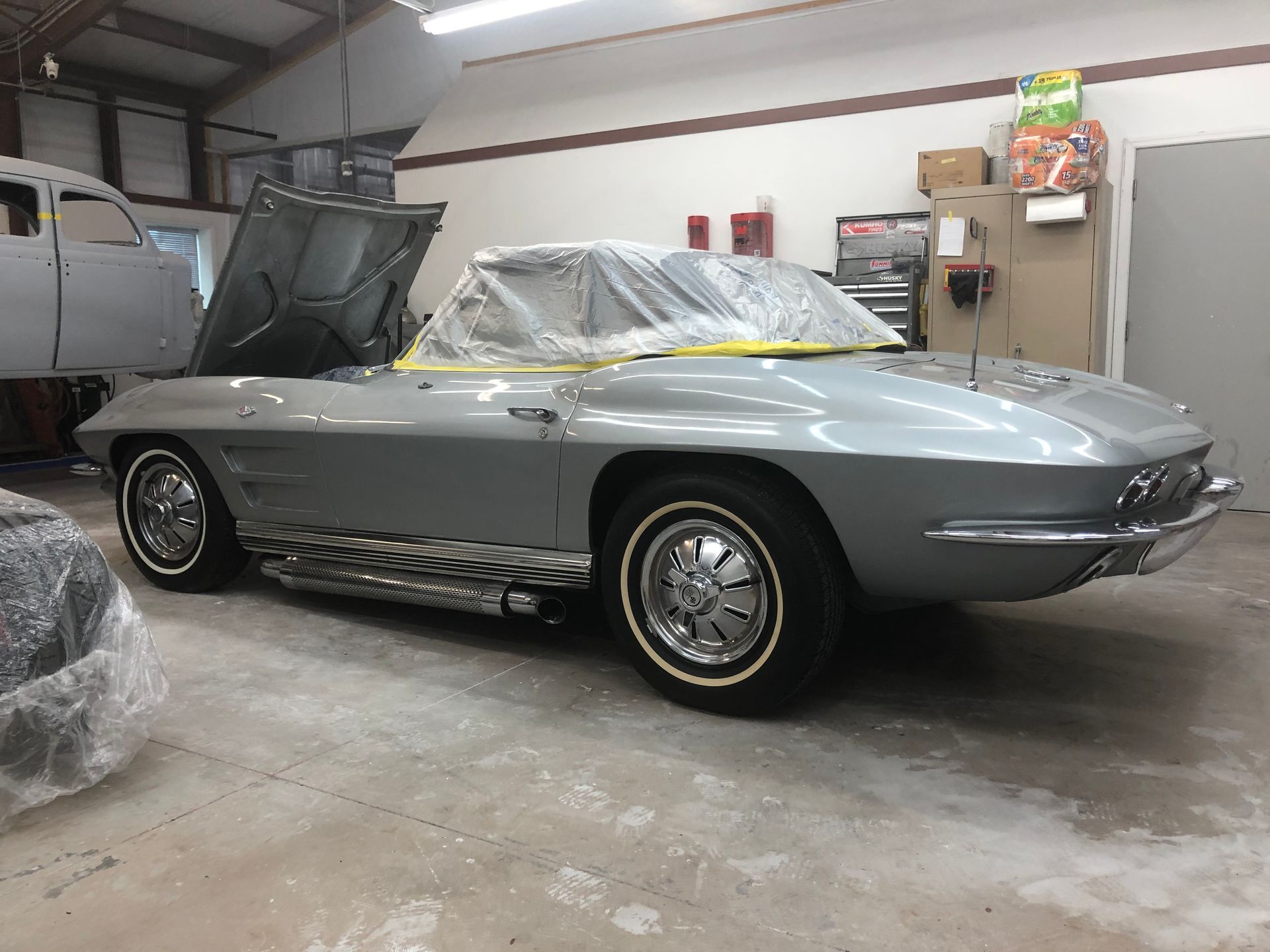 A silver classic Chevrolet Corvette sits in a garage with its hood raised and the windshield covered in protective plastic.