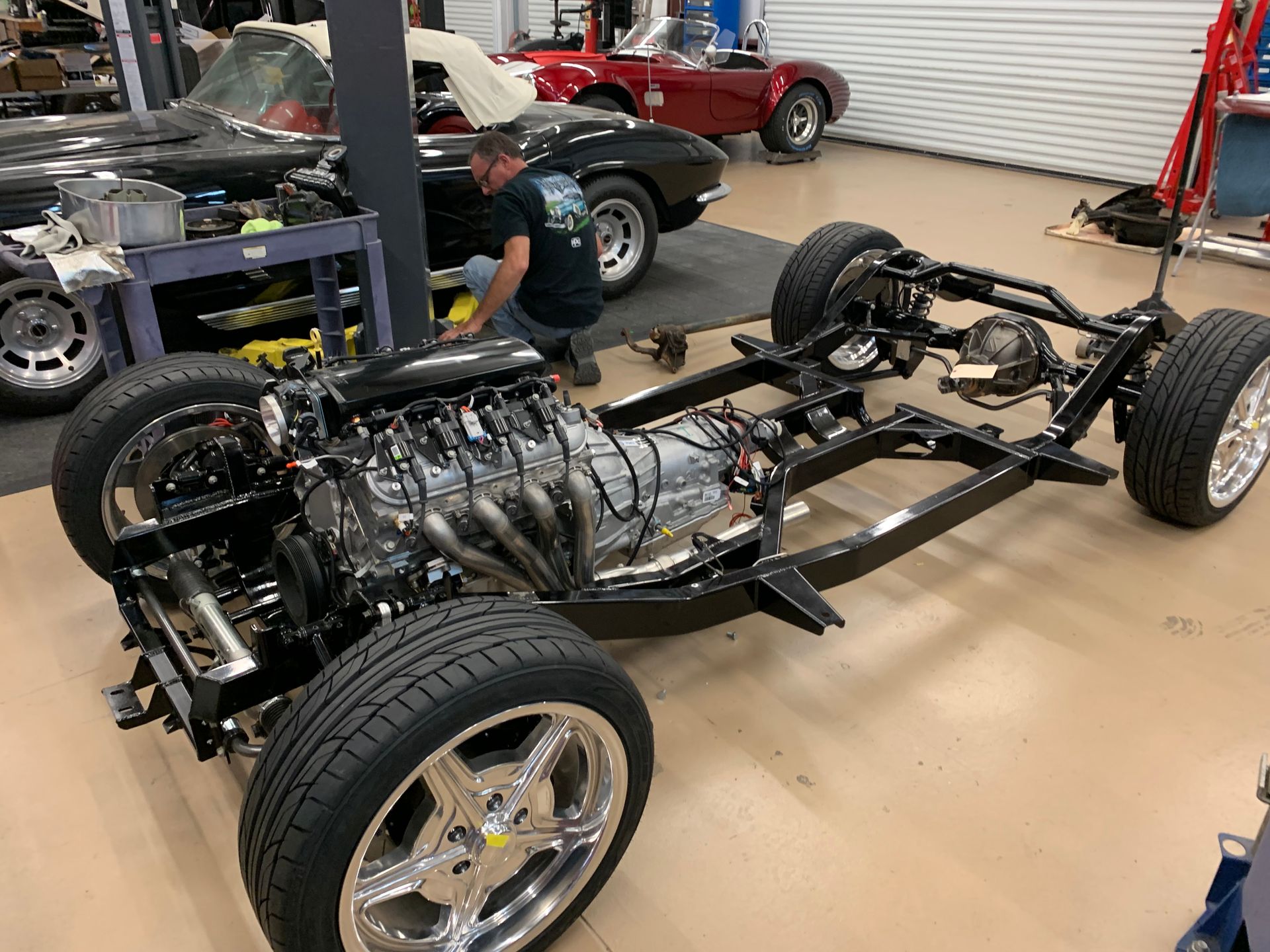 A mechanic works on a classic car chassis with an installed engine and new wheels in a workshop.