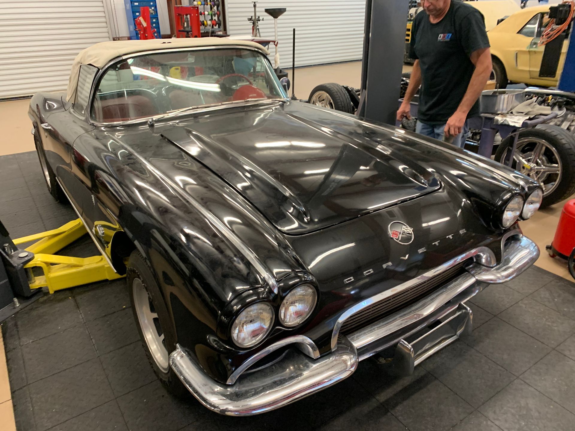 A black vintage Chevrolet Corvette convertible parked in a garage next to a person and car parts.