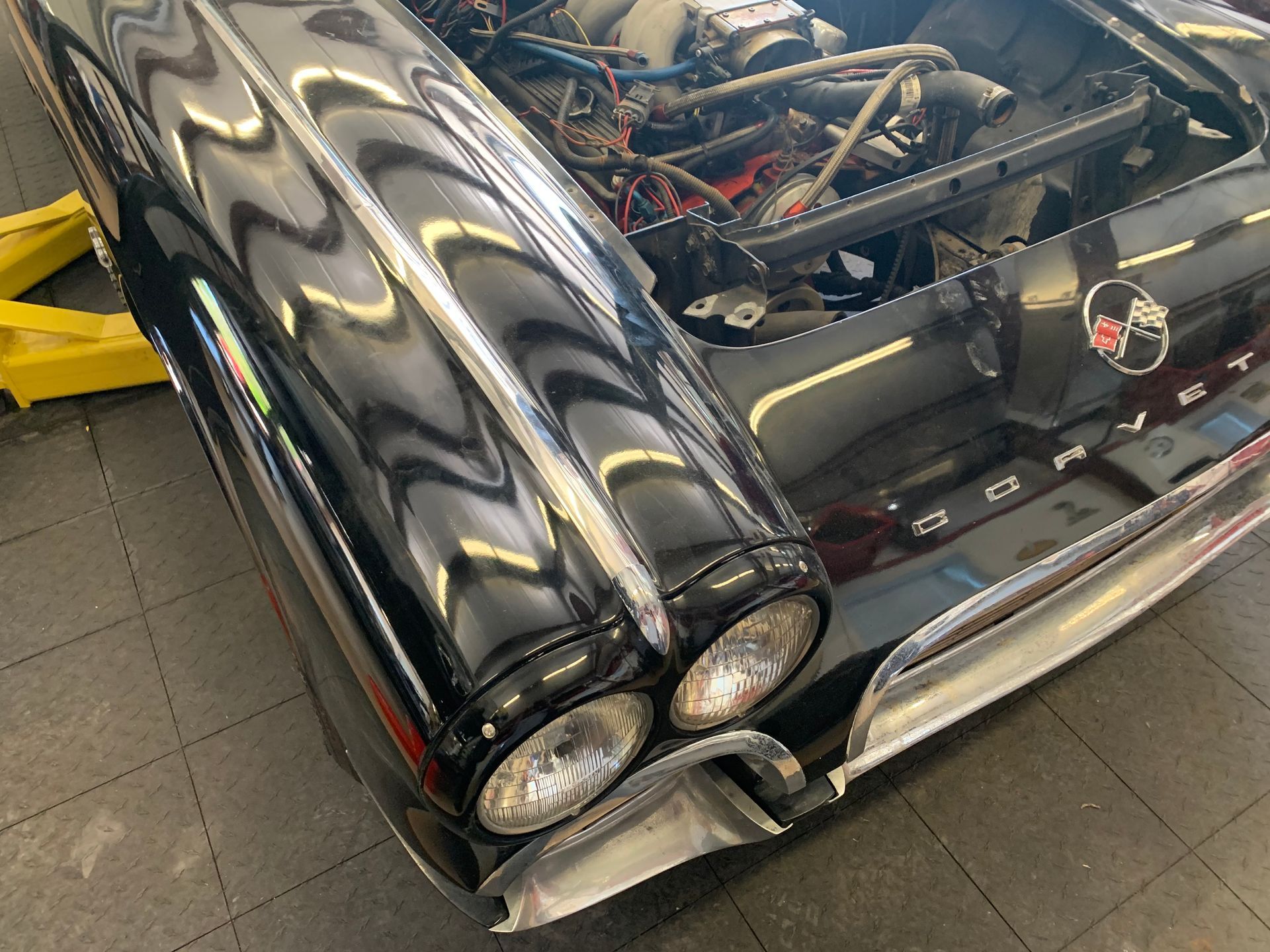 A black vintage Corvette undergoing maintenance in a garage, featuring prominent dual headlights and chrome accents.