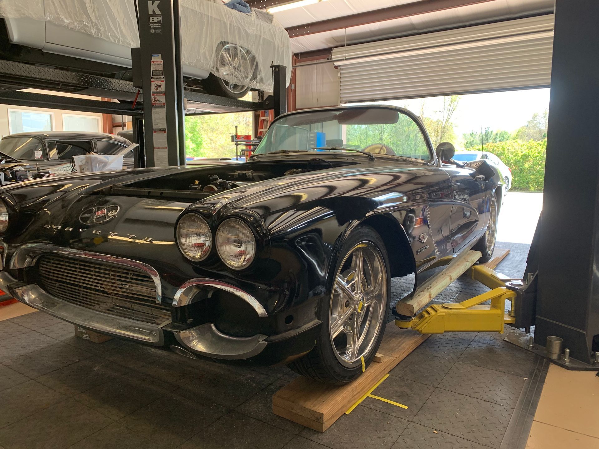 A black vintage Corvette is raised on a yellow automotive lift inside a garage, with another car visible above it.