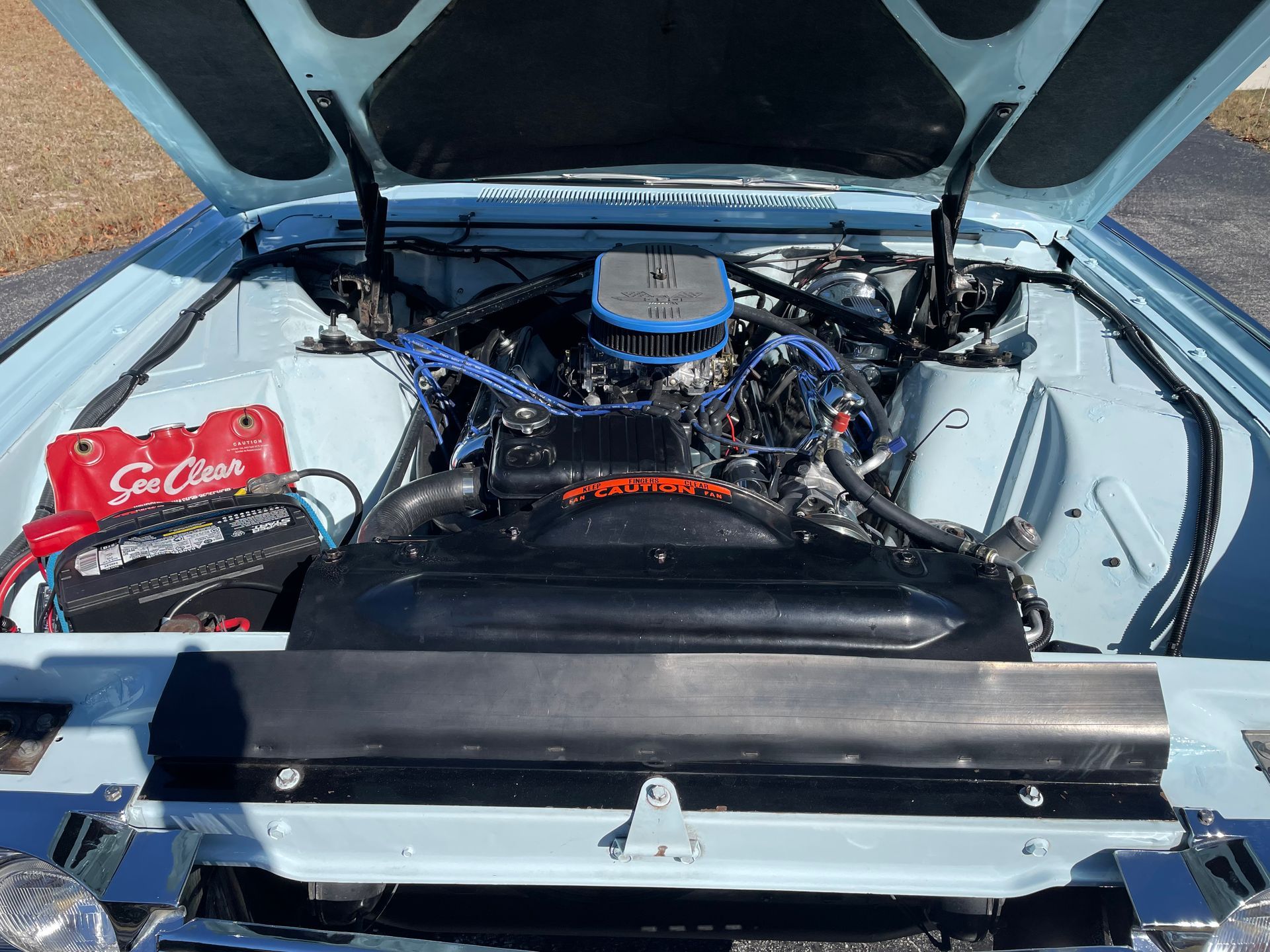 The open hood of a pale blue vintage car reveals a detailed engine bay with a black radiator shroud and red battery.