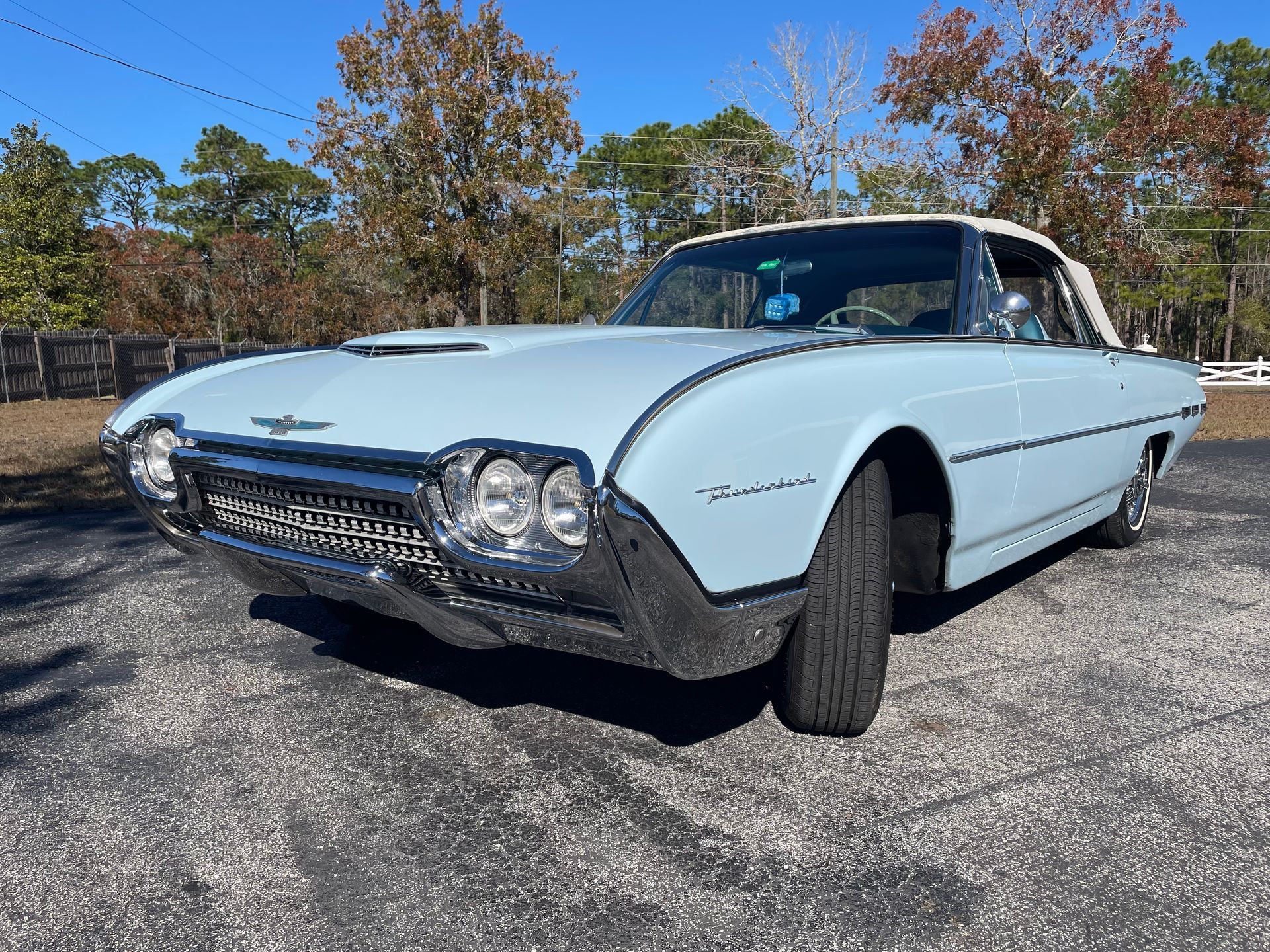 A light blue vintage Ford Thunderbird convertible parked on an asphalt lot with trees in the background under a blue sky.