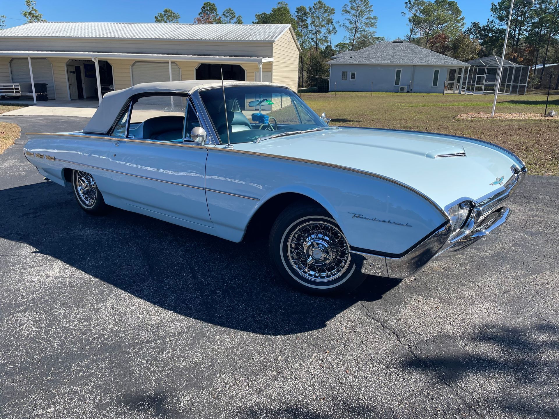 A vintage light blue Ford Thunderbird convertible parked on an asphalt driveway in front of suburban houses.