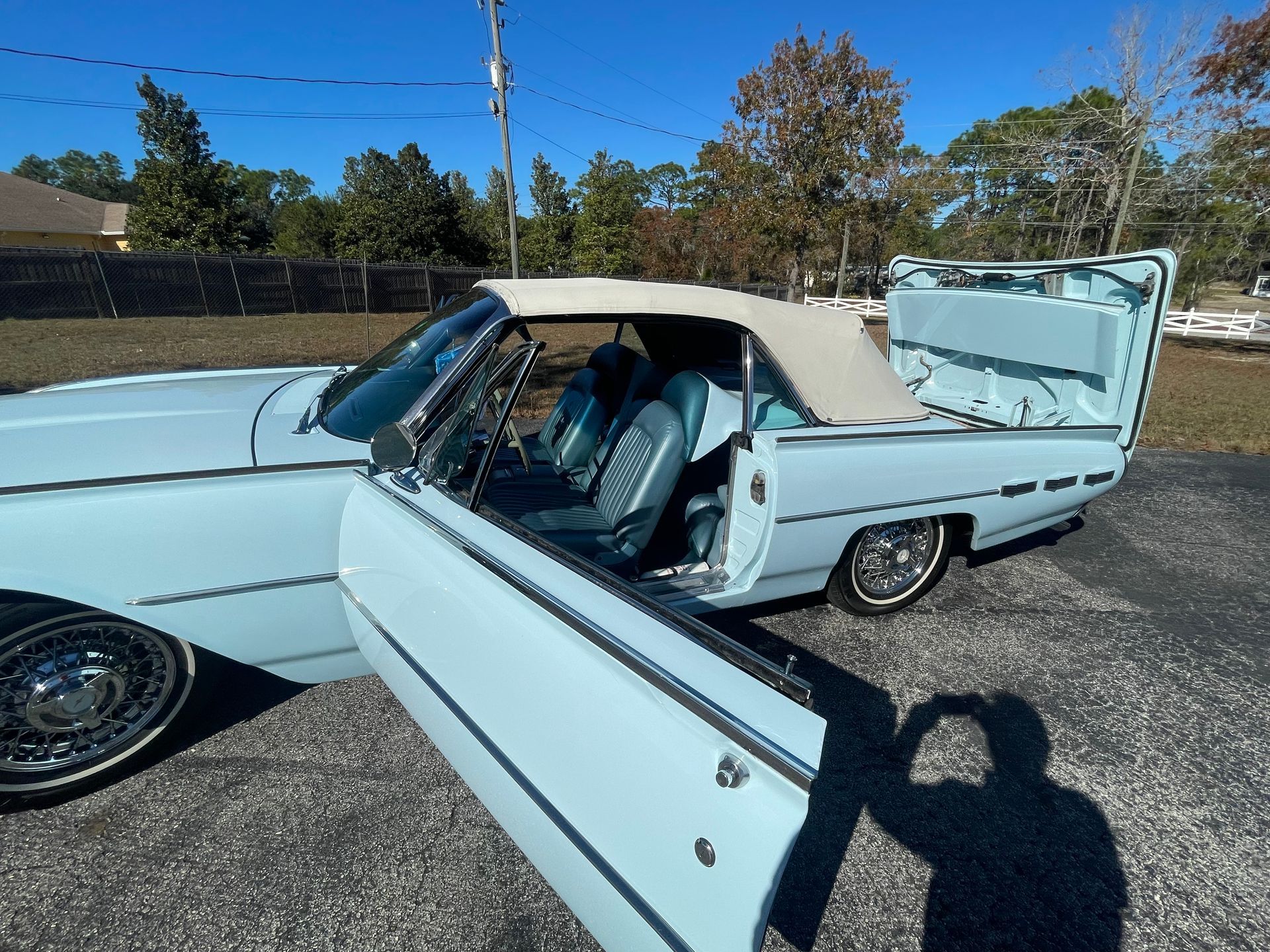 A light blue vintage convertible with its top down and trunk open, parked on an asphalt lot under a clear blue sky.