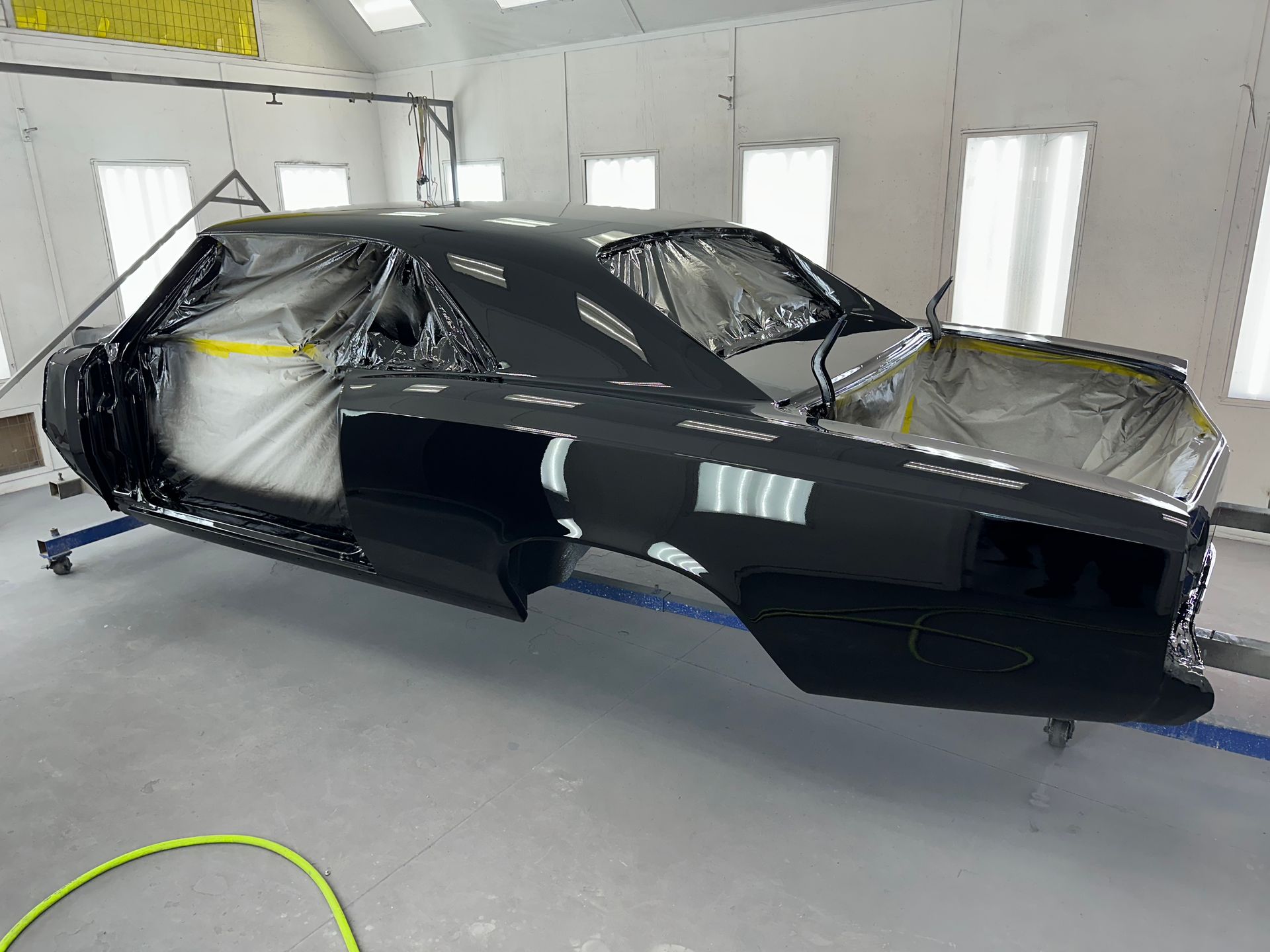A black vintage muscle car body shell masked for painting, mounted on a stand inside a professional auto body shop.