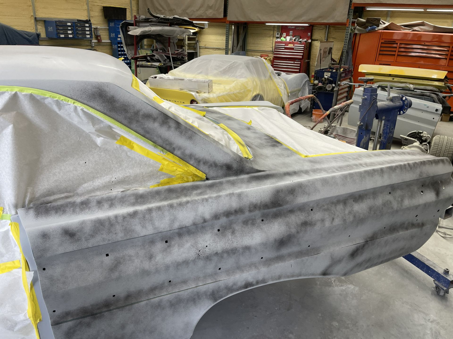 A car quarter panel in a workshop, covered in gray body filler primer and partially masked with yellow tape.
