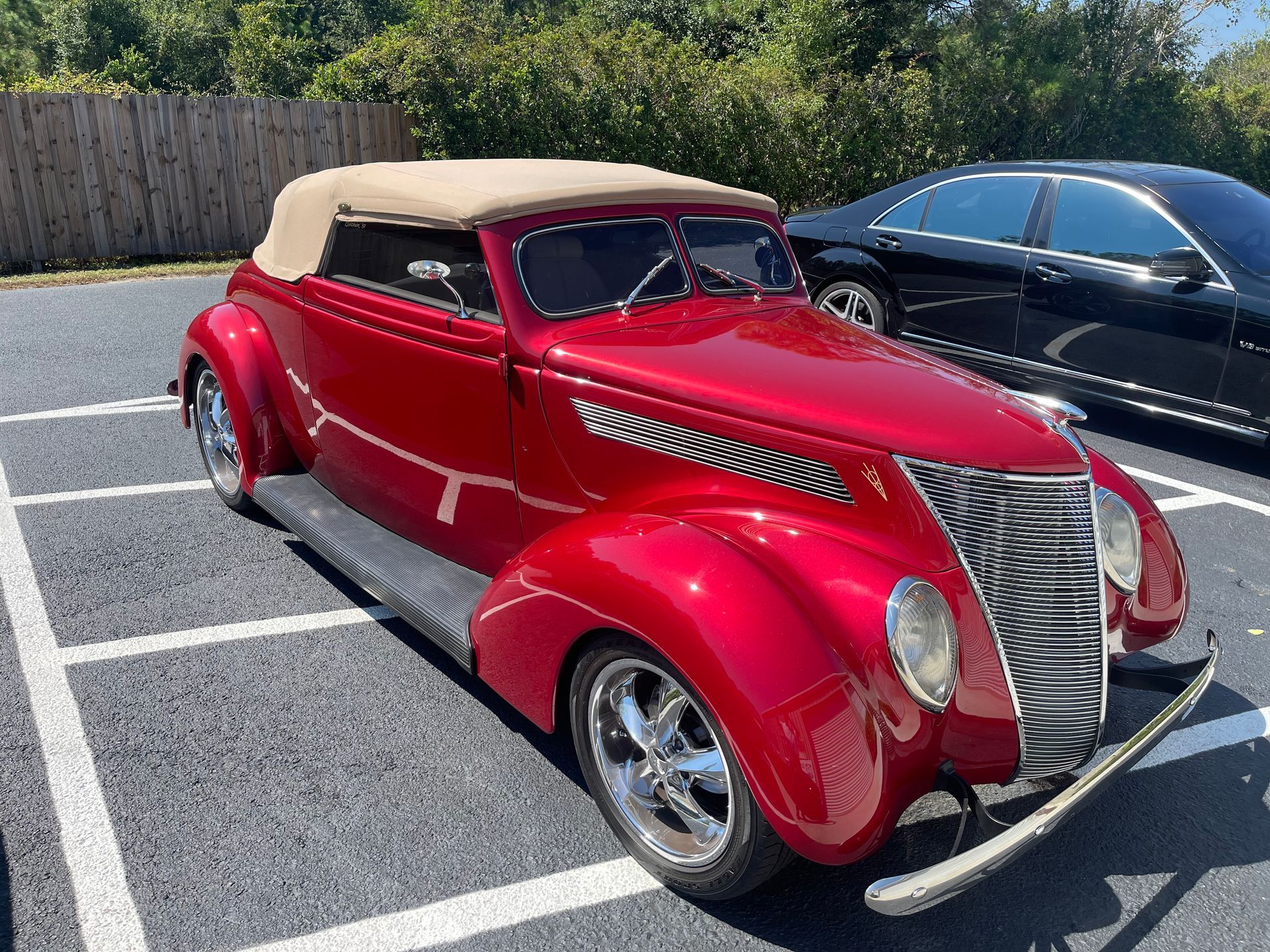 A bright red vintage Ford roadster with a tan convertible top parked in a parking lot next to a black car.