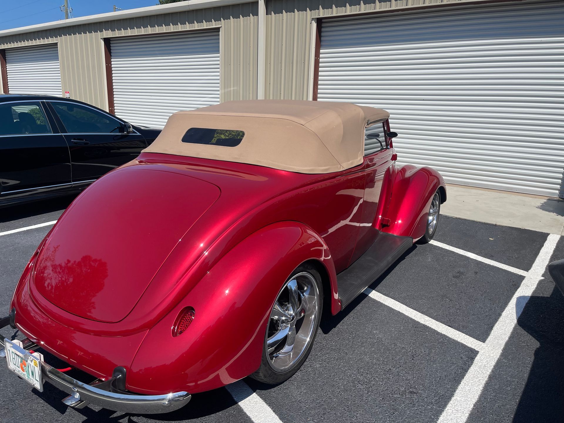 A candy-apple red vintage convertible with a tan soft top parked in a paved lot next to a black sedan.