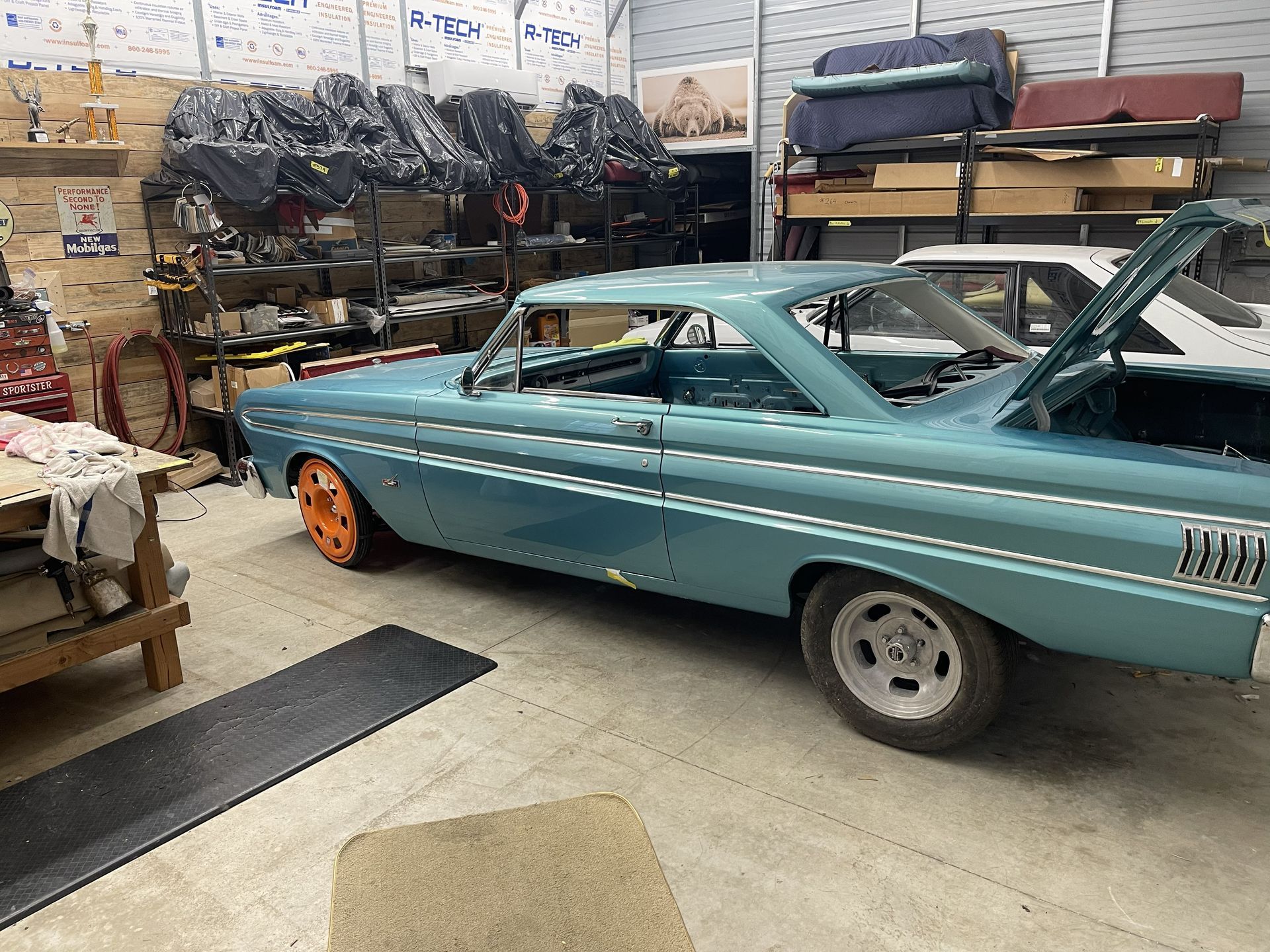 A light blue vintage Ford Falcon with its trunk open, parked inside a garage filled with car parts and storage shelving.