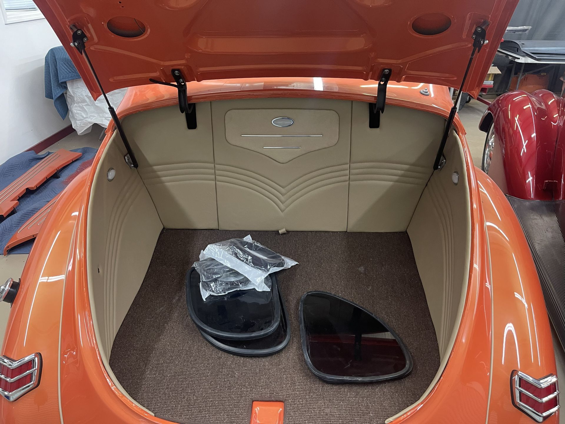 Open trunk of an orange classic car showing a custom beige interior, brown carpeting, and a few loose car parts.