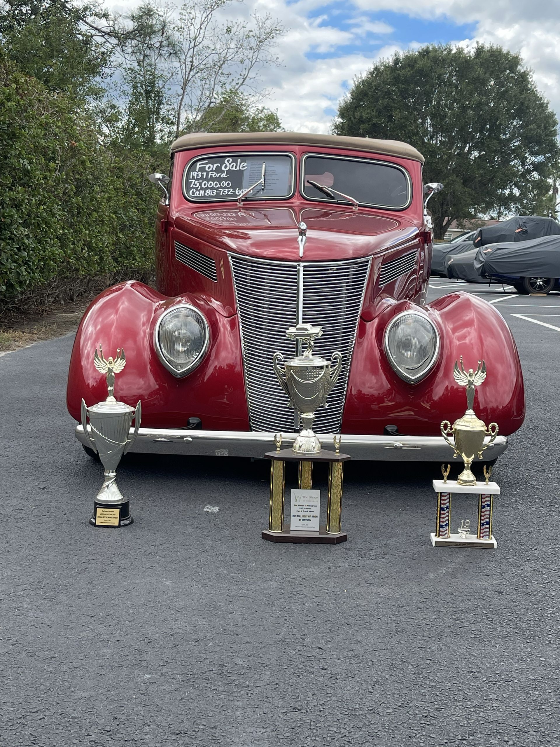 A cherry-red classic vintage car parked outdoors with three gold trophy cups placed on the ground in front of it.