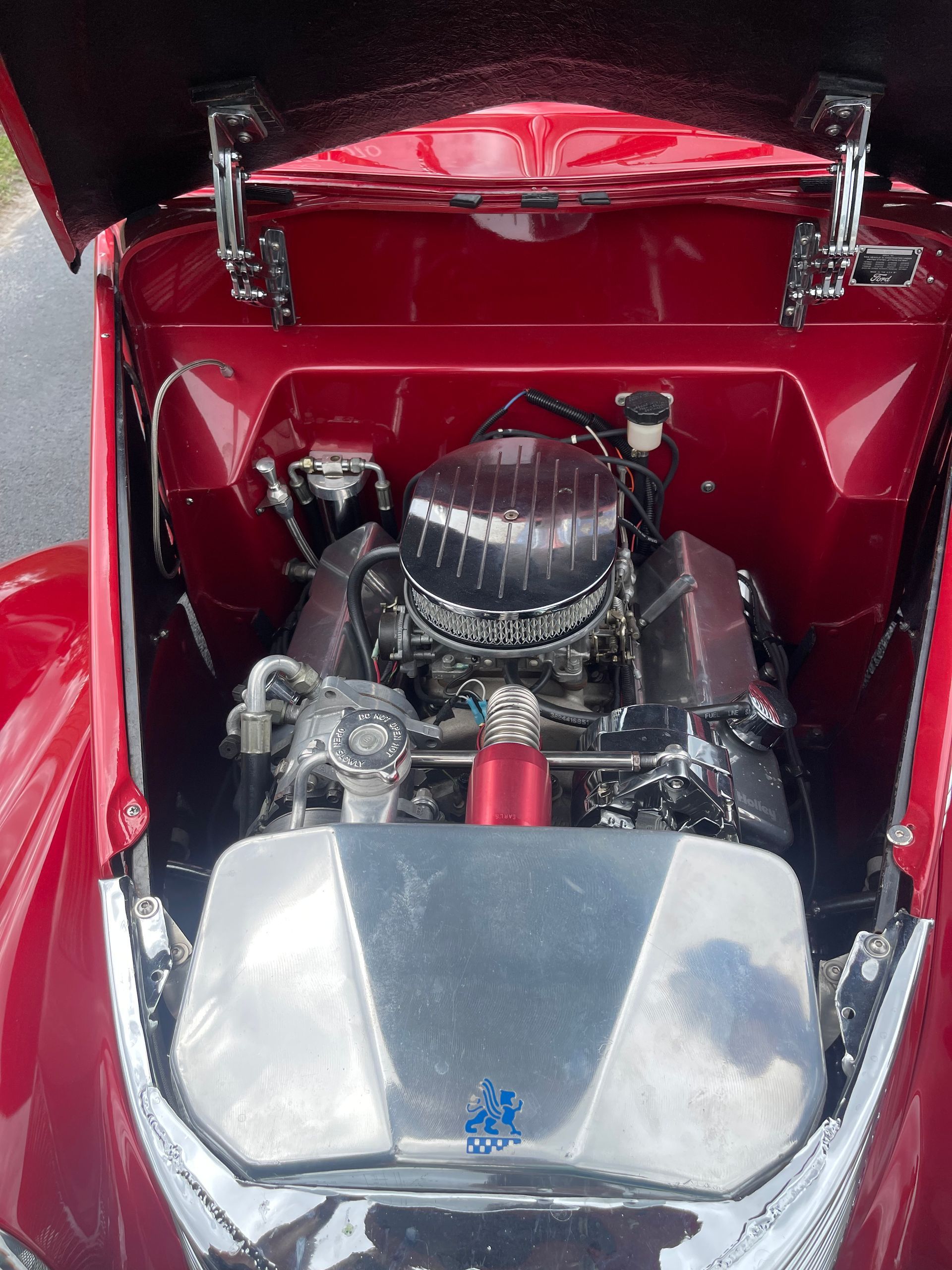 The open hood of a bright red car revealing a clean, chrome-accented V8 engine compartment.