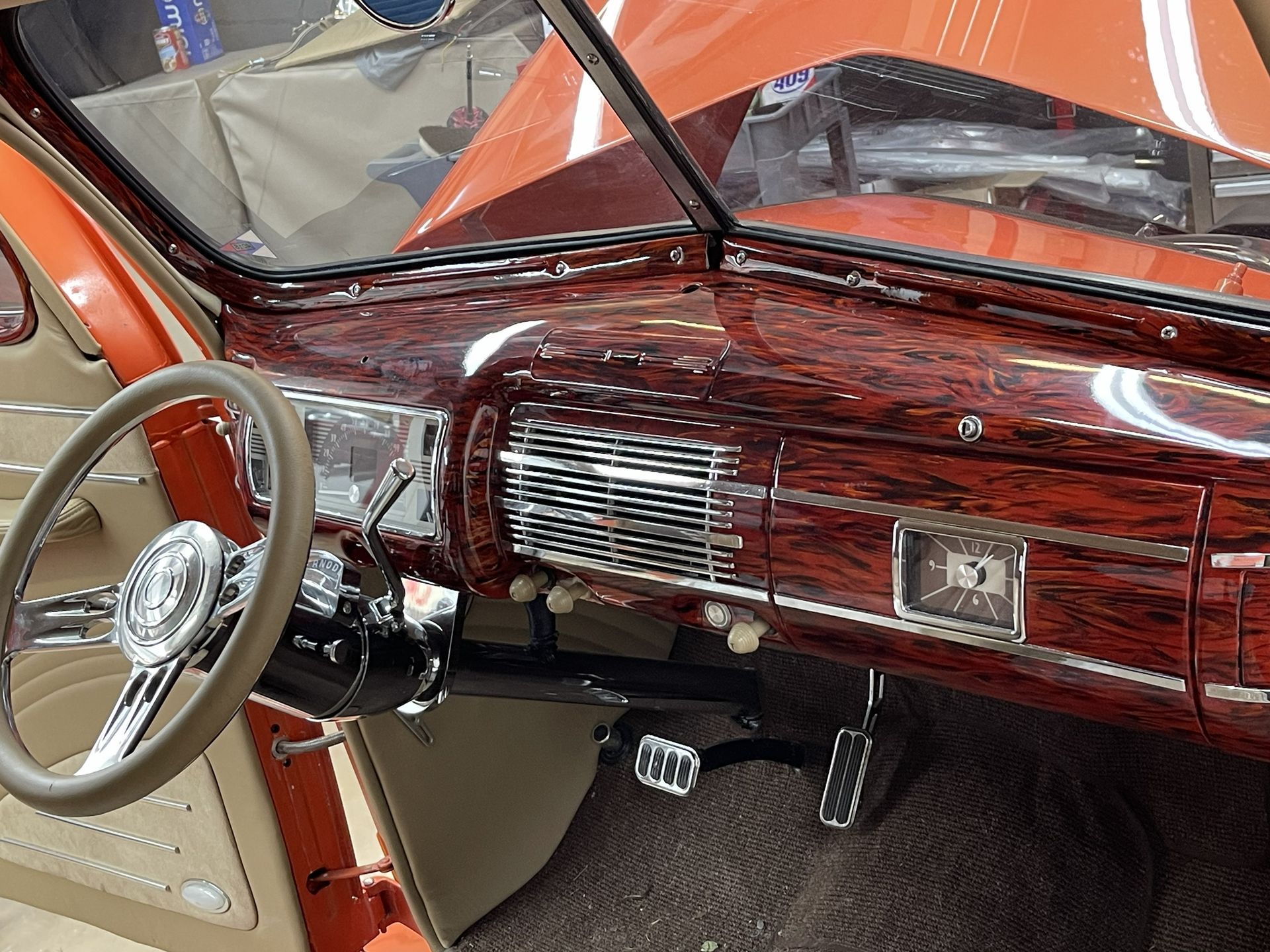 The interior of a classic car featuring a polished, wood-grain patterned dashboard, a beige steering wheel, and pedals.