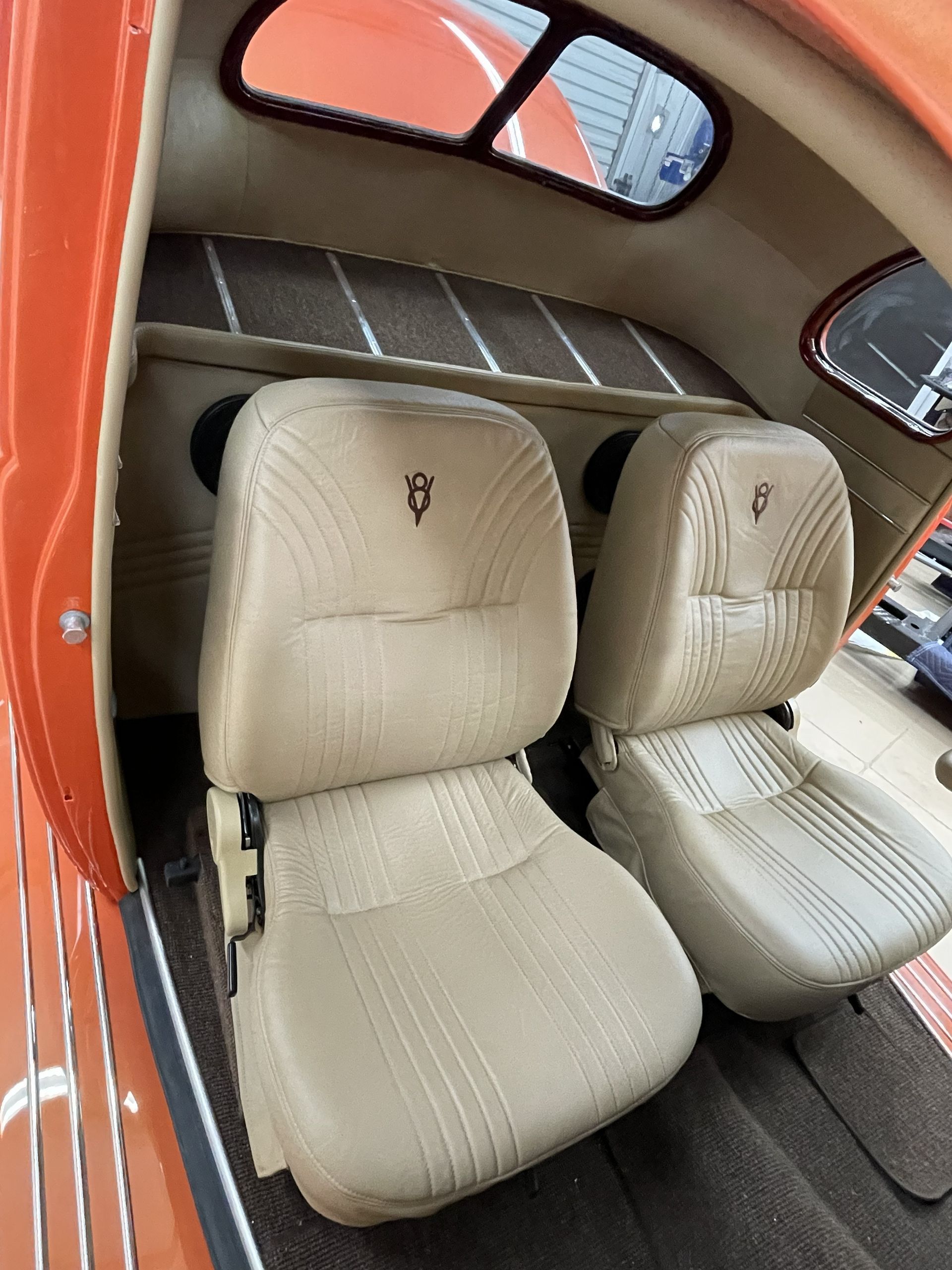 The interior of an orange vintage vehicle features two beige bucket seats with custom emblems on a gray carpeted floor.
