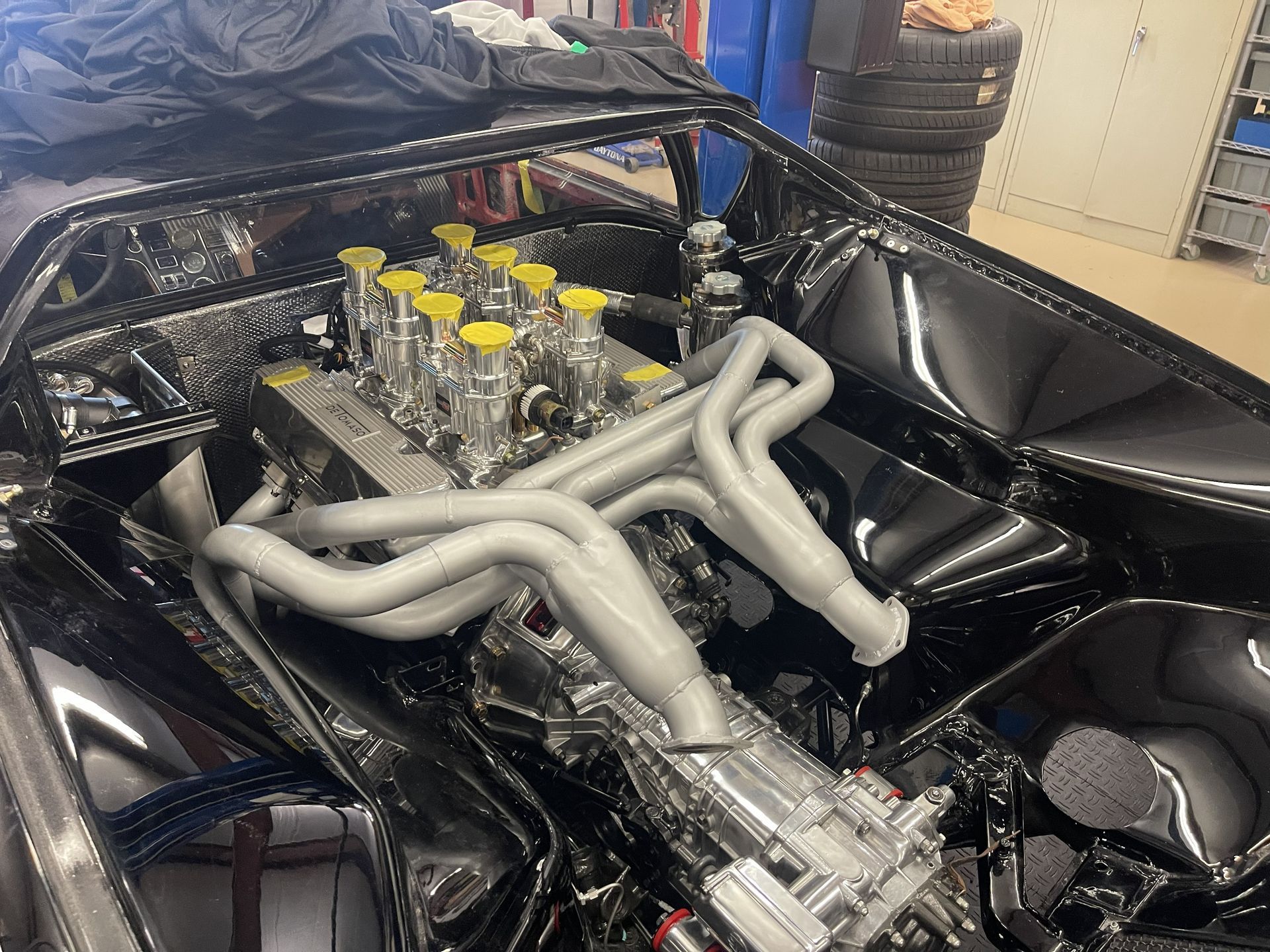 A high-performance V8 engine with silver exhaust headers and yellow intake covers mounted in a black car chassis.