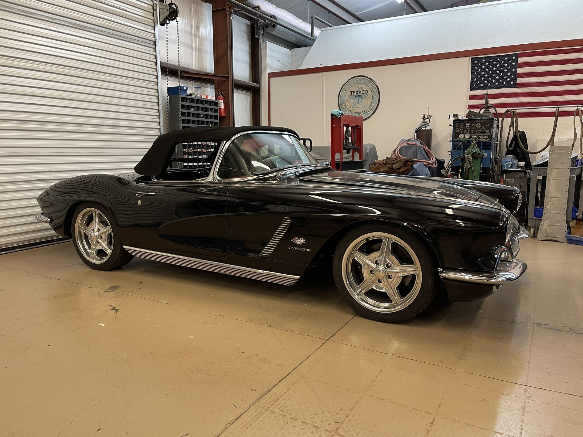 A black classic convertible Chevrolet Corvette parked inside a workshop with a U.S. flag on the wall.