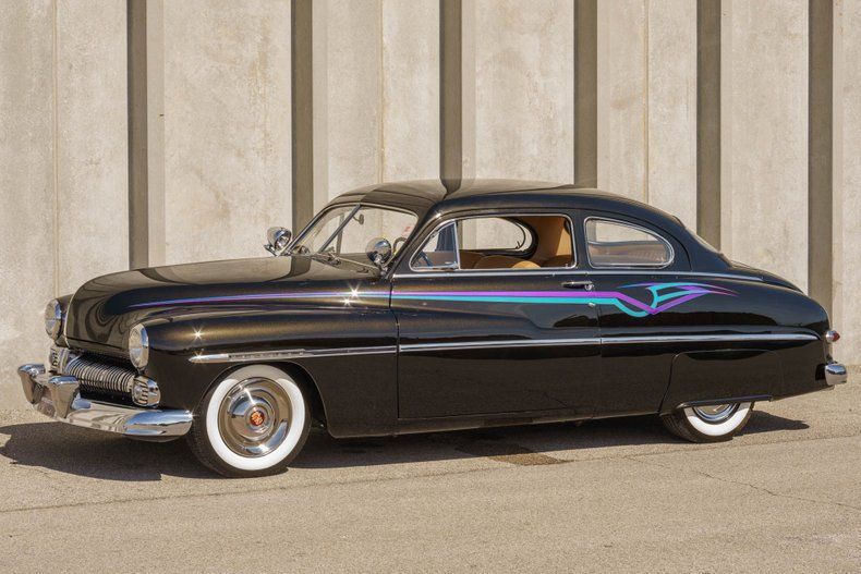 A customized vintage black coupe with white-wall tires and teal and purple pinstriping parked against a concrete wall.