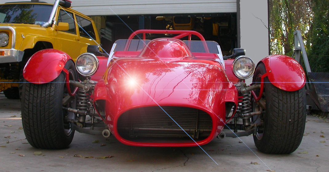 A bright red, open-wheel sports car with round headlights, parked outside a garage next to a yellow vehicle.