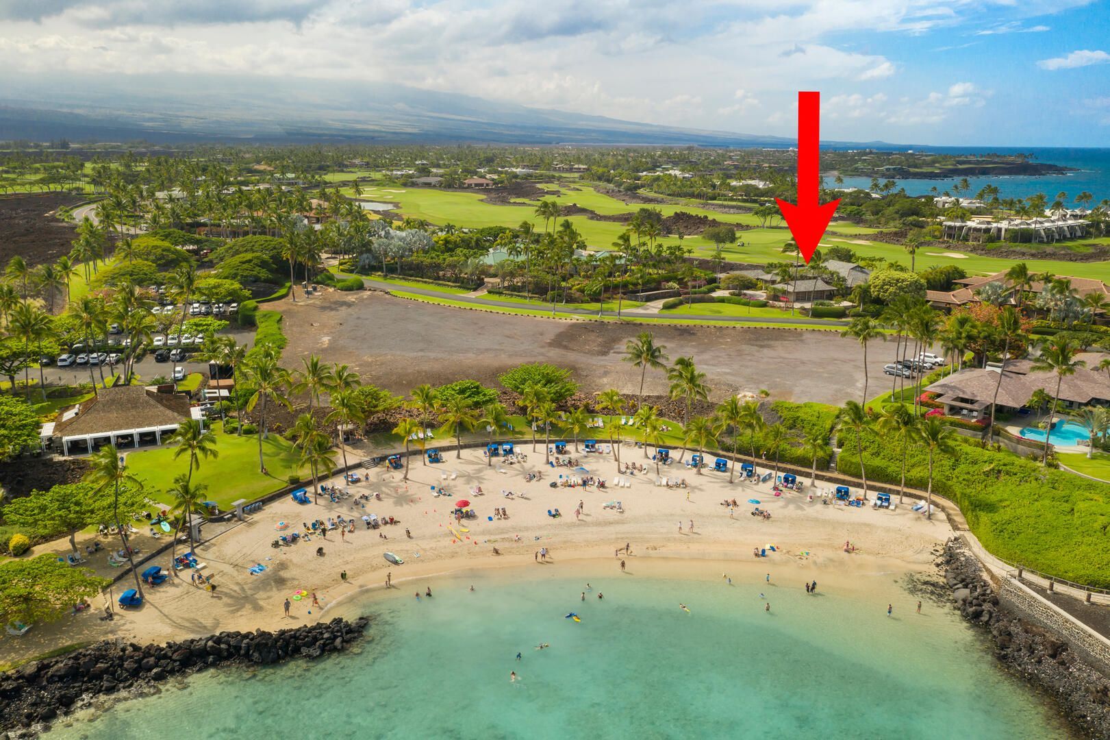 Aerial view of a sandy beach with people, turquoise water, and a resort. Arrow points to a building.