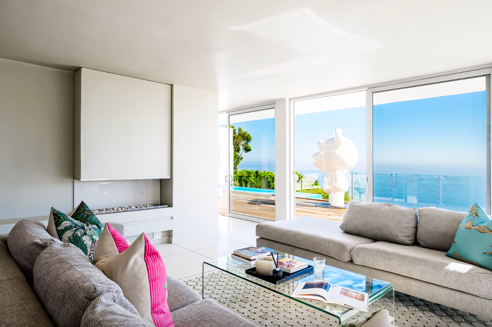 Modern living room with ocean view; gray sofas, glass coffee table, white walls, and bright blue sky.