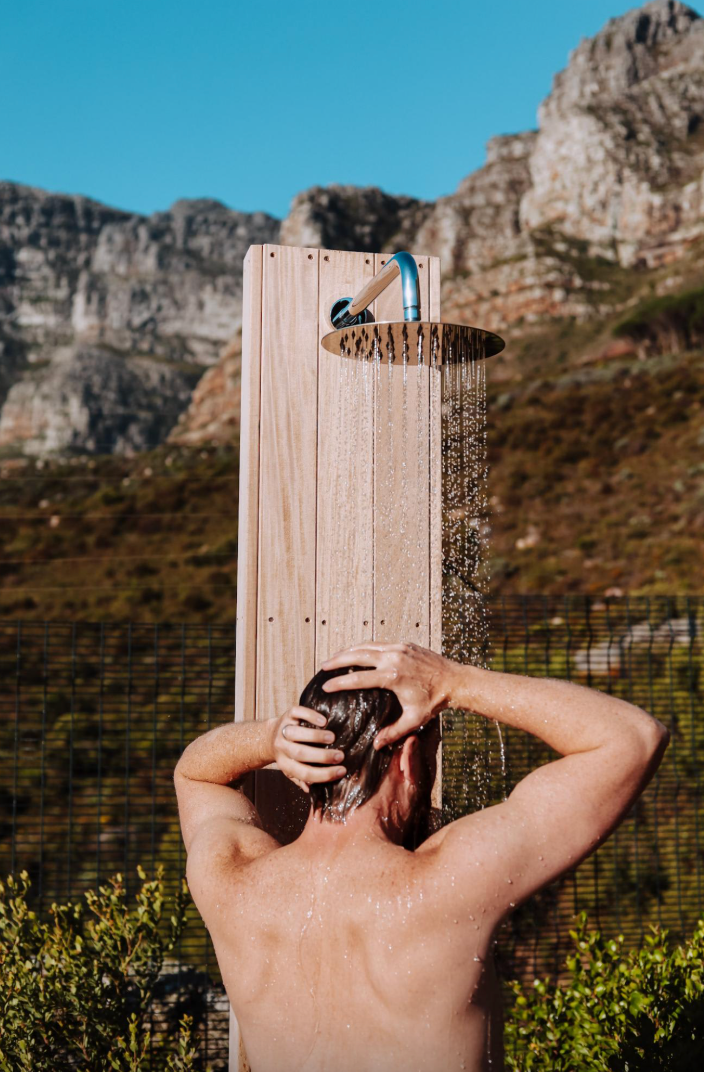 Person showering outdoors with a mountain backdrop. Water cascades from shower head onto their head.