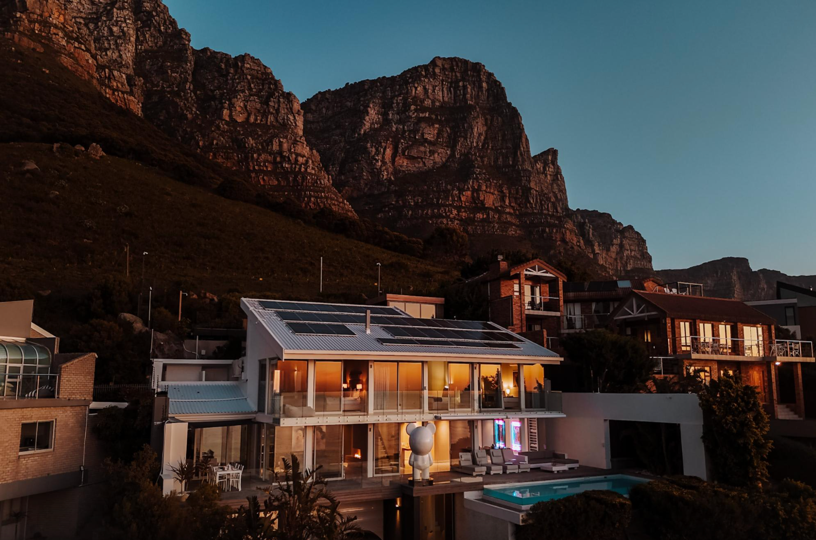 Modern white house with illuminated interior, pool, and mountain backdrop at sunset.