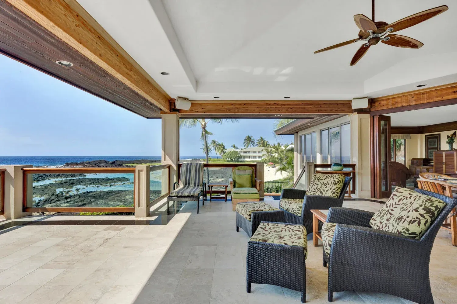 Patio with ocean view, wicker furniture, light stone floor, brown ceiling beams, blue sky.