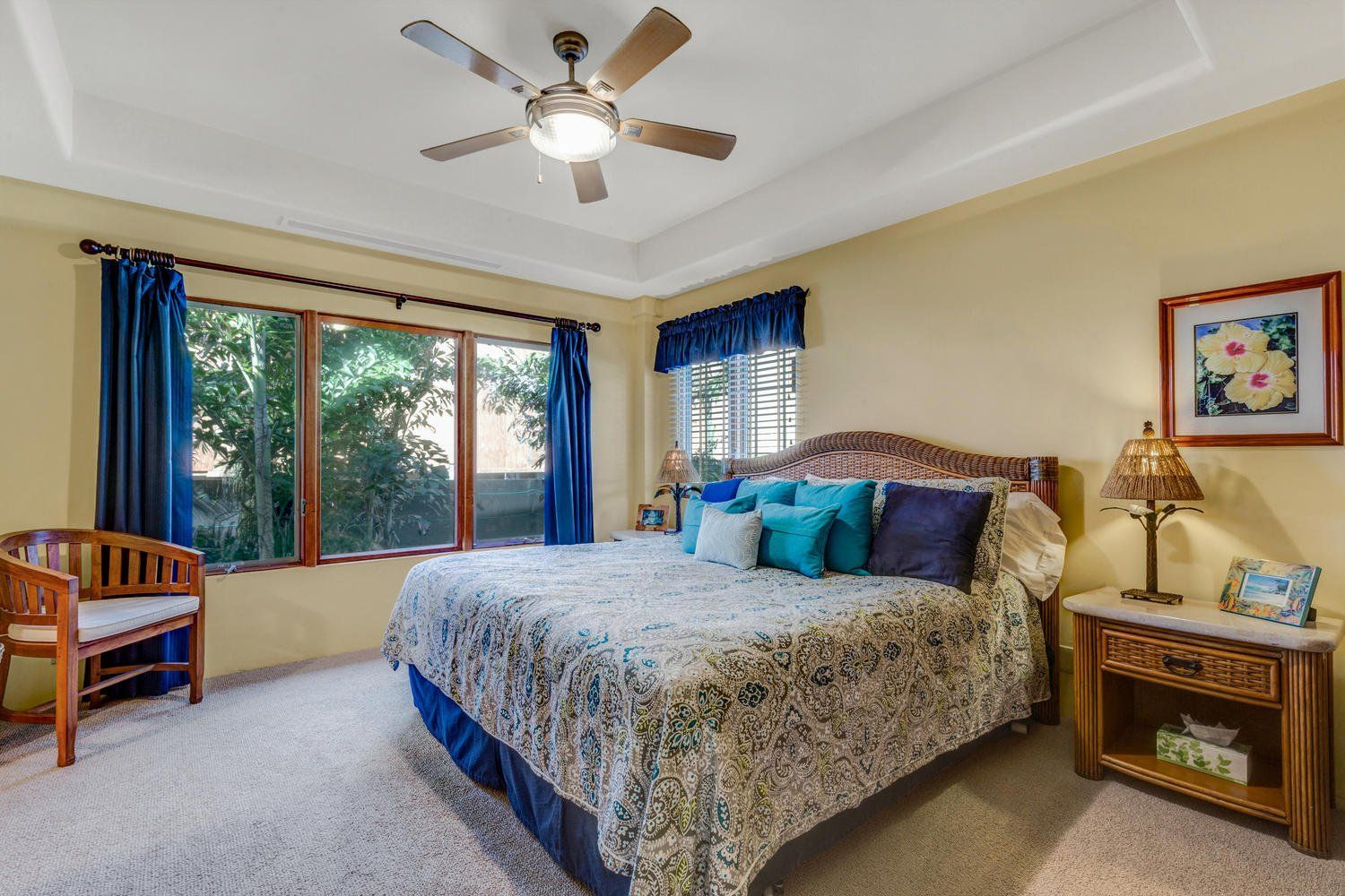 Bedroom with a bed, armchair, and window with blue curtains. Yellow walls, beige carpet, and a ceiling fan.