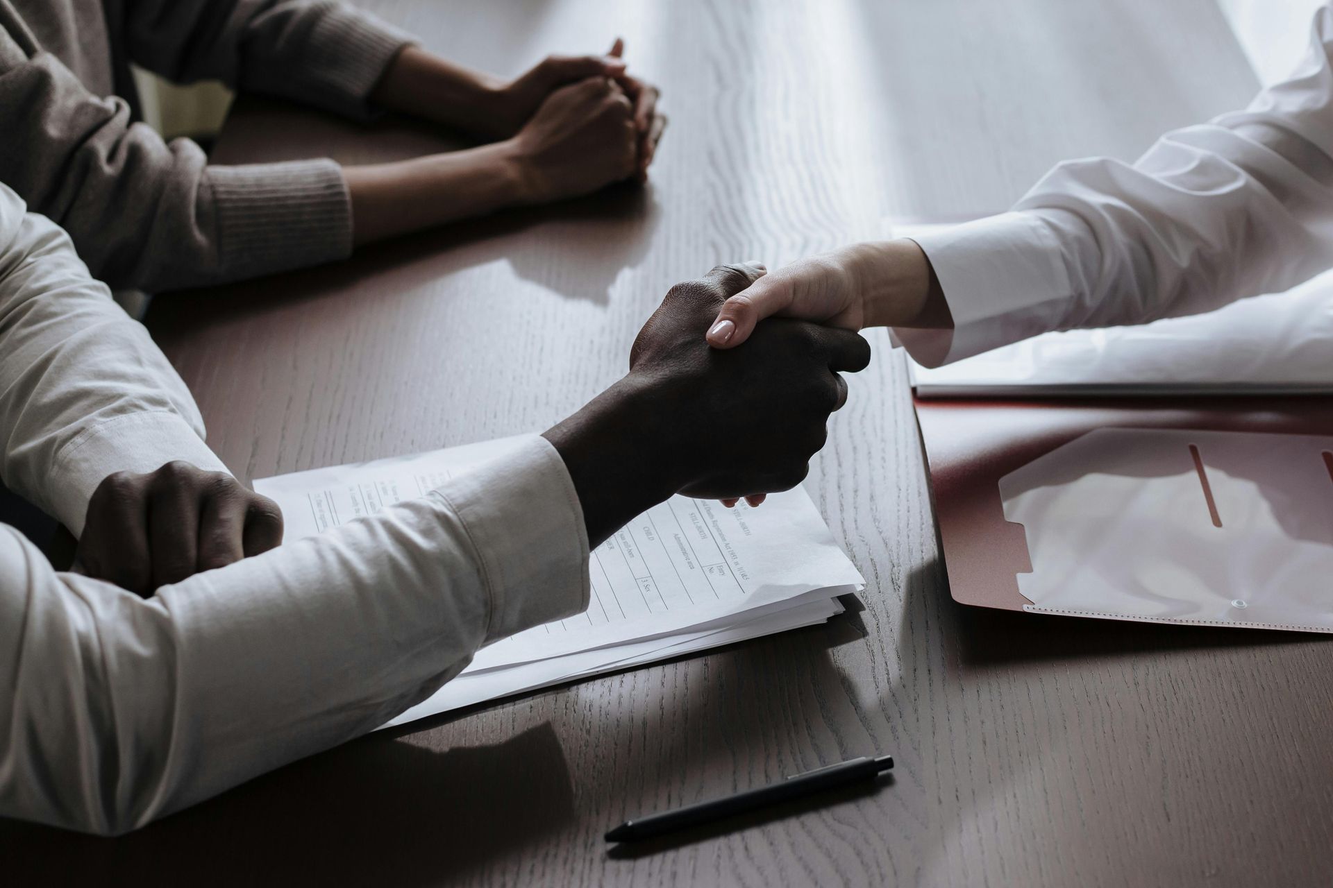 Two people shaking hands over a wooden table, paperwork and a pen visible.T