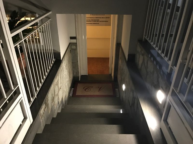 Una scala poco illuminata conduce a una porta illuminata, con ringhiere in metallo e applique a parete di notte.