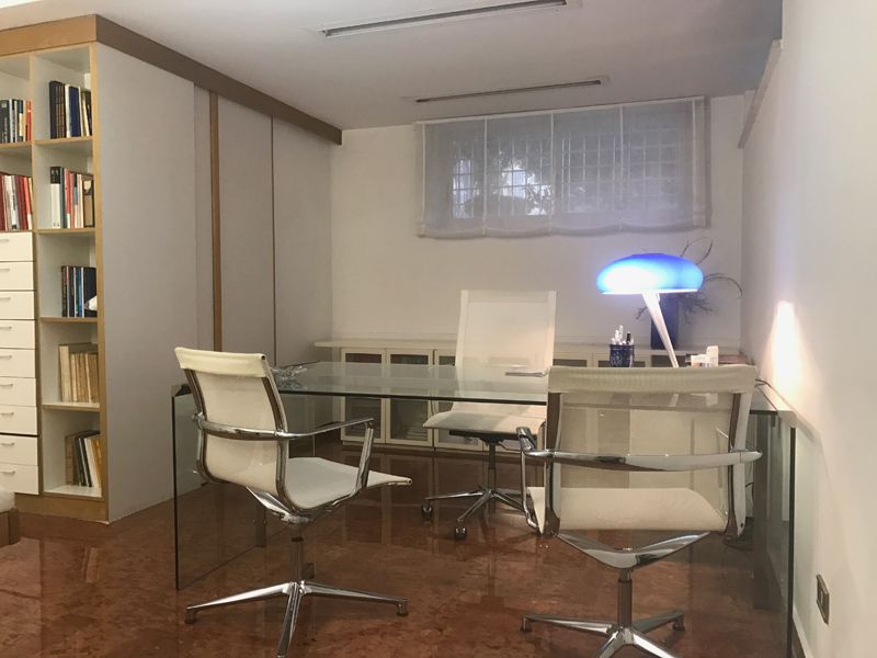Moderno ufficio domestico con scrivania in vetro, sedie bianche, librerie e lampada blu.