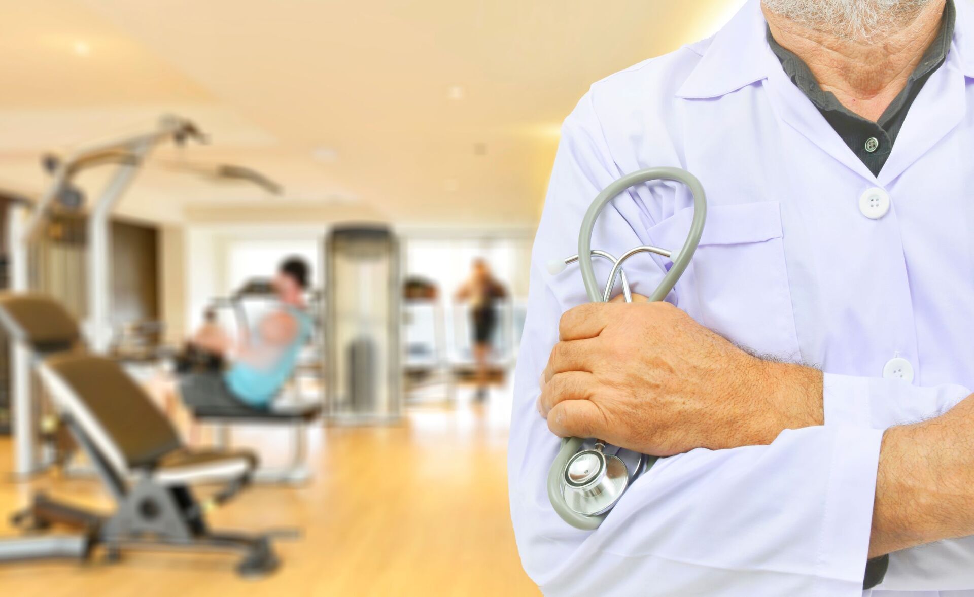 Un medico in camice bianco tiene in mano uno stetoscopio in una luminosa palestra d'ospedale, con attrezzi ginnici sullo sfondo.