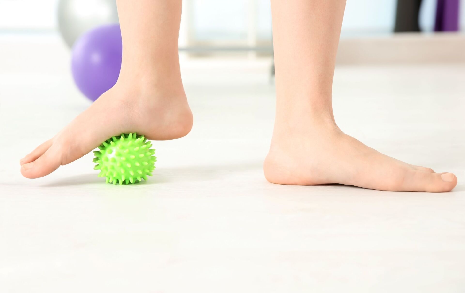 Piedi che premono su una palla da massaggio verde e appuntita su un pavimento bianco, con una palla viola sullo sfondo.