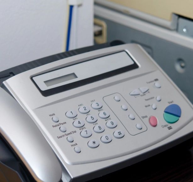 Silver fax machine with number pad and display, paper in feed tray.