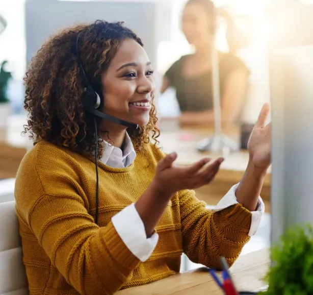 Woman wearing headset, smiling while gesturing at computer in office setting.